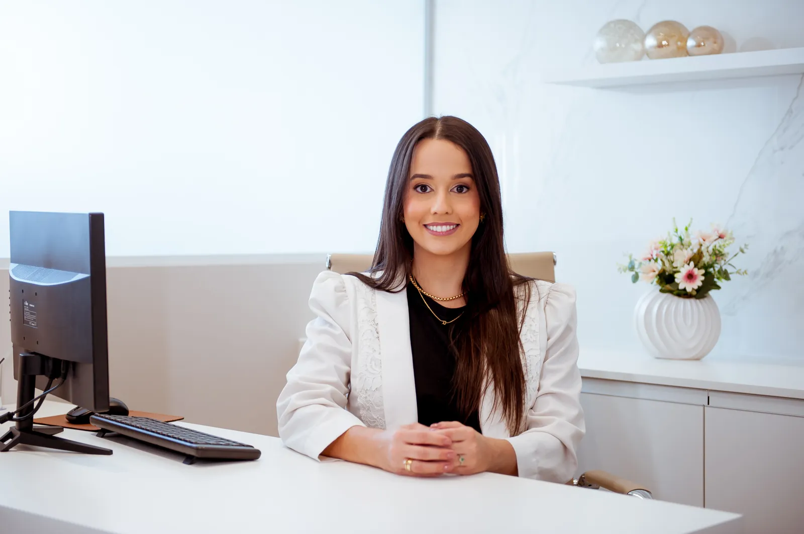Dra. Raissa Amaral sentada à mesa do consultório claro, com flores e prateleira ao fundo