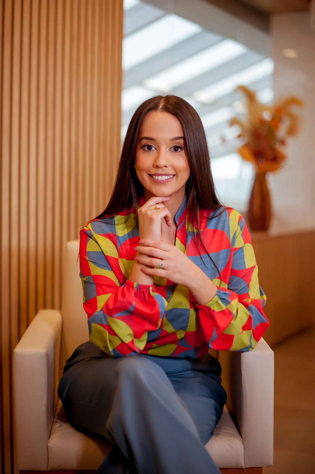 Dra. Raissa Amaral sentada em uma poltrona clara, sorrindo, com blusa estampada colorida